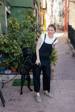 Load image into Gallery viewer, Woman in the Paloma linen black jumpsuit with adjustable straps, playfully interacting with a black dog in a vibrant street setting.