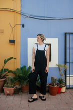 Load image into Gallery viewer, Woman wearing the Paloma linen black jumpsuit with adjustable straps, standing against a colorful wall with plants around.