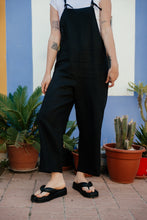 Load image into Gallery viewer, Close-up of the Paloma linen black jumpsuit, featuring adjustable straps, worn by a woman with stylish black sandals in a colorful outdoor setting.
