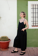Load image into Gallery viewer, Model in the Women's Black Linen Jumpsuit Vega – Relaxed Fit Overall with Adjustable Straps, standing in a casual pose by the green wall, emphasizing the loose fit and summer vibe.