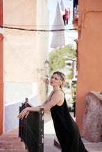 Load image into Gallery viewer, Woman in the Women's Black Linen Jumpsuit Vega leaning on a fence, surrounded by vibrant architecture, emphasizing the stylish wide-leg design.