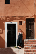 Load image into Gallery viewer, Woman in a Women's Black Linen Jumpsuit Vega standing casually against a colorful wall, showcasing the relaxed fit and adjustable straps of the overall.