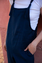 Load image into Gallery viewer, Close-up of a woman wearing a dark blue Linen jumpsuit with adjustable straps, showcasing pocket details against a pink background.