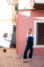 Load image into Gallery viewer, Full body shot of a woman in a dark blue Linen jumpsuit with adjustable straps, posing casually in a colorful street scene.