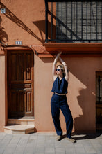 Load image into Gallery viewer, Model posing with raised arms while wearing Sofa Killer dark blue linen trousers Lucia and a matching crop top against a vibrant wall.
