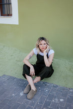 Load image into Gallery viewer, Woman seated casually in a linen dark brown color jumpsuit with adjustable straps Paloma, leaning against a green wall.