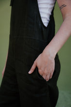 Load image into Gallery viewer, A woman's hand holding the pocket of her linen dark brown color jumpsuit with adjustable straps Paloma, highlighting its functional style.