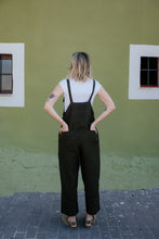 Load image into Gallery viewer, Woman from behind in the Linen dark brown color jumpsuit with adjustable straps Paloma, accentuating the wide-leg style against a green wall.