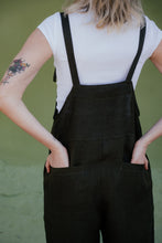 Load image into Gallery viewer, Back view of a woman wearing the Linen dark brown color jumpsuit with adjustable straps Paloma, showcasing its pockets and relaxed fit.