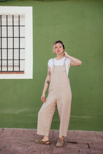 Load image into Gallery viewer, Model wearing a loose linen beige jumpsuit with adjustable straps, styled casually with a white tee against a vibrant green backdrop.