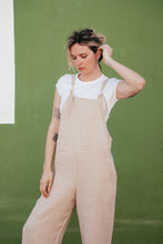 Load image into Gallery viewer, Model wearing a loose linen beige color jumpsuit with adjustable straps, styled casually against a green backdrop.