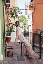 Load image into Gallery viewer, Model in a loose linen beige color jumpsuit with adjustable straps, relaxing in a vibrant street setting surrounded by plants.