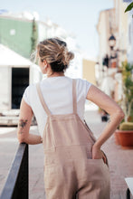 Load image into Gallery viewer, Back view of a model wearing a loose linen beige color jumpsuit with adjustable straps, showing off its relaxed fit and pockets.