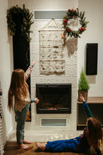 Load image into Gallery viewer, Sofa Killer tricolor unisex kids onesie Groove, featuring two children pointing at a festive wall calendar near a cozy fireplace, highlighting the comfort and style of the onesie.