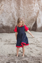 Load image into Gallery viewer, Girl wearing the Sofa Killer linen dark blue dress with red details, playfully standing on a sandy beach.