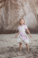 Load image into Gallery viewer, A young girl wearing a Sofa Killer linen beige dress with violet color details, posing joyfully on a sandy beach near a rocky backdrop.