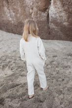 Load image into Gallery viewer, A child wearing a Sofa Killer linen beige unisex kids onesie, standing on a sandy surface with a rocky background. Perfect for spring and summer adventures.