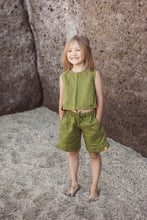 Load image into Gallery viewer, A smiling girl standing in sand, dressed in the Sofa Killer linen moss green unisex set with shorts, perfect for family gatherings and casual outings.