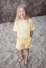 Load image into Gallery viewer, A happy child wearing the Sofa Killer linen yellow unisex set with shorts, smiling at the camera while standing on a sandy surface. The outfit features a short-sleeve top and comfortable shorts, perfect for warm weather.