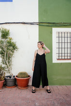 Load image into Gallery viewer, Women's Black Linen Jumpsuit Vega – Relaxed Fit Overall with Adjustable Straps, model posing against a green wall, showcasing the comfortable design and elegant style.
