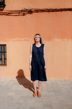 Load image into Gallery viewer, Model wearing a Women's dark blue linen sleeveless dress Costa Blanca by SofaKiller, standing in front of a textured orange wall. The button-down summer dress features a gathered waist, showcasing a relaxed and stylish look perfect for warm weather.
