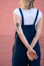 Load image into Gallery viewer, Back view of a woman in a dark blue Linen jumpsuit with adjustable straps, highlighting her tattoos and the relaxed fit at a pink wall.
