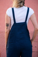 Load image into Gallery viewer, Rear view of a woman in a dark blue Linen jumpsuit with adjustable straps, focusing on the design and pockets, set against a pink backdrop.
