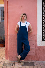 Load image into Gallery viewer, Model wearing a Linen dark blue color jumpsuit with adjustable straps Paloma, standing against a pink wall, showcasing a relaxed and stylish look.
