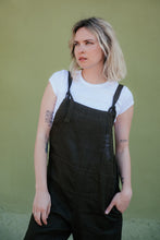 Load image into Gallery viewer, Close-up of a woman in a linen dark brown color jumpsuit with adjustable straps Paloma, highlighting her stylish pose against a green background.
