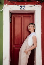 Load image into Gallery viewer, Fashionable shot of a model in a beige linen jumpsuit with adjustable straps, leaning against a red door, showcasing effortless style.

