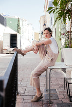 Load image into Gallery viewer, Model showcasing a loose linen beige color jumpsuit with adjustable straps, sitting on a table and striking a pose.
