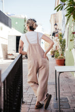 Load image into Gallery viewer, Model from behind in a loose linen beige color jumpsuit with adjustable straps, standing casually in an outdoor setting.
