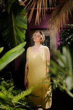 Load image into Gallery viewer, A woman wearing the Women's yellow linen sleeveless dress Marisol by SofaKiller, standing in a lush garden surrounded by vibrant green plants, showcasing the dress's cheerful color and casual style.
