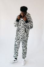 Load image into Gallery viewer, Full view of a man wearing the Sofa Killer animal men onesie zebra. The cozy outfit showcases a striking black and white zebra design, complemented by casual white sneakers.
