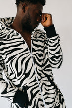 Load image into Gallery viewer, Stylish Sofa Killer animal men onesie in zebra pattern, featuring a hood and black cuffs. Model posing thoughtfully with one hand to his face.
