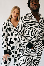 Load image into Gallery viewer, A man and a woman posing in matching Sofa Killer animal onesie designs, featuring Zebra and cow patterns.
