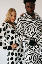 Load image into Gallery viewer, Casual pose of a man and woman in Sofa Killer animal men onesies, one in zebra and the other in cow print.
