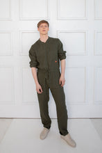 Load image into Gallery viewer, Sofa Killer dark green linen men onesie, model wearing it while standing confidently against a minimalistic backdrop.
