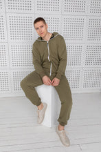 Load image into Gallery viewer, Model wearing Sofa Killer olive color onesie, casually seated on a pedestal, showcasing comfort and style.
