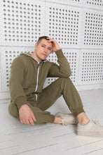Load image into Gallery viewer, Model in Sofa Killer olive color onesie, sitting on the floor with a thoughtful pose, highlighting the cozy fit of the outfit.

