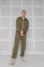 Load image into Gallery viewer, Model standing confidently in Sofa Killer olive color onesie, with a minimalist backdrop, emphasizing the trendy design.
