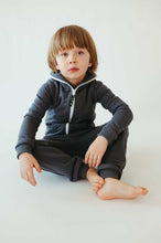 Load image into Gallery viewer, Sofa Killer unisex kids onesie Rock worn by a young boy sitting cross-legged on a white background, showcasing the dark grey color and white zipper.
