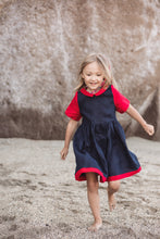Load image into Gallery viewer, A cheerful young girl running on the beach in a Sofa Killer linen dark blue dress with red details. The soft fabric gives her freedom to move and play, perfect for a fun day out.
