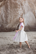 Load image into Gallery viewer, A close-up view of a little girl in a Sofa Killer linen beige dress with violet color details, playfully walking on the beach, showcasing the dress's design.
