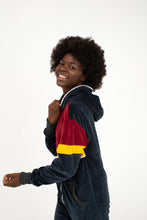 Load image into Gallery viewer, Model smiling while wearing the Sofa Killer women velour onesie Kansas, showcasing the vibrant maroon and yellow accents against a dark grey background.
