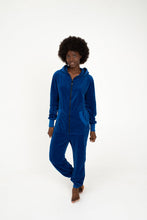 Load image into Gallery viewer, Woman in a Sofa Killer royal blue velour onesie Nevada, standing with hands in pockets, radiating casual elegance.
